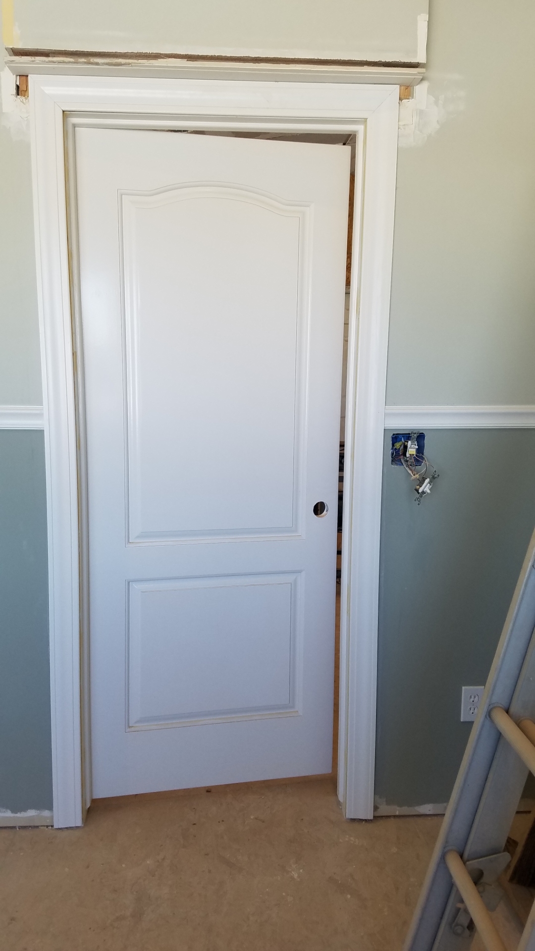 A white solid wood door with a fresh coat of paint hung in a door frame in a room with two tone green and gray paint with a chair rail and molding around the door. No door knob installed